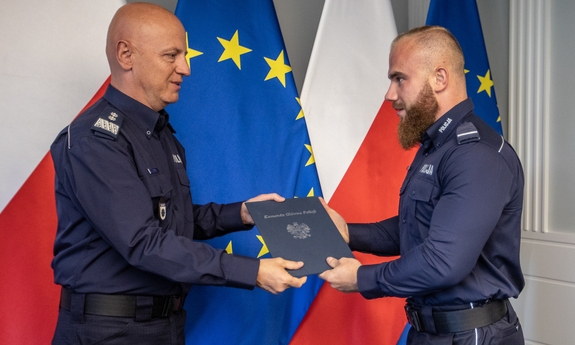 Komendant Główny Policji gen. insp. Jarosław Szymczyk z policjantem na tle flag Polski i Unii Europejskiej, na ścianie wisi godło Polski.