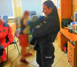 policjantka karmiąca butelka noworodka owiniętego w policyjna bluzę. W tle widać ratownika medycznego i zamazaną graficznie młodą kobietę - matkę dziecka