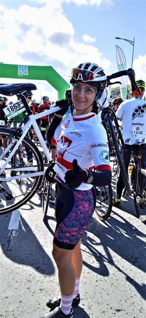 Kobieta w stroju sportowym jako zawodniczka, trzyma na ramieniu rower, stojąc  przed metą