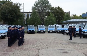 W tle stoją nowe, oznakowane radiowozy. Na pierwszym planie stoją policjanci, do których przemawia Zastępca Komendanta Głównego Policji nadinsp. Roman Kuster