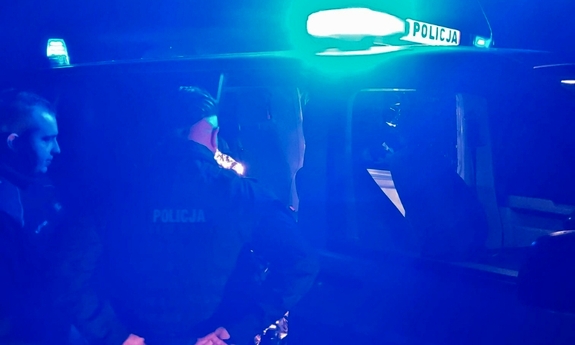 pora nocna, policjanci stoją przy radiowozie, który ma włączone sygnały świetlne