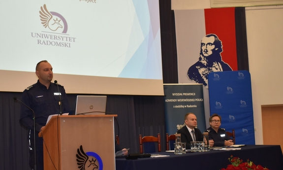 policjant stoi przy mównicy, obok niego przy stole siedzi mężczyzna i policjantka