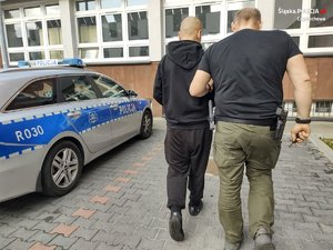 nieumundurowany policjant doprowadza zatrzymanego, w tle policyjny radiowóz