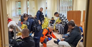 uchodźcy w pomieszczeniu siedzą na krzesłach, część z nich okryta jest kocami, policjanci udzielają im pomocy