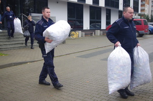 policjanci noszą worki z nakrętkami w tle budynek  i zaparkowane auta