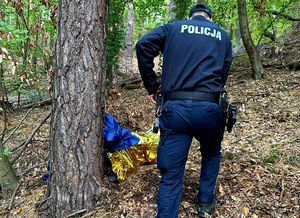 Policjant stoi przy leżącym pod drzewem zaginionym mężczyźnie. Mężczyzna jest przykryty kocem ratunkowym.