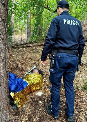 Policjant stoi przy leżącym pod drzewem zaginionym mężczyźnie. Mężczyzna jest przykryty kocem ratunkowym.