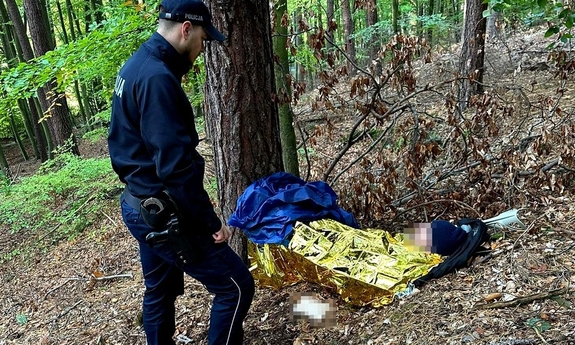 Policjant stoi przy leżącym pod drzewem zaginionym mężczyźnie. Mężczyzna jest przykryty kocem ratunkowym.