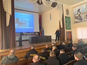 Policjant, który szkoli funkcjonariuszy stoi na scenie przed ekranem, na którym wyświetlana jest prezentacja. Przed sceną w rzędach na fotelach siedzą uczestnicy szkolenia.