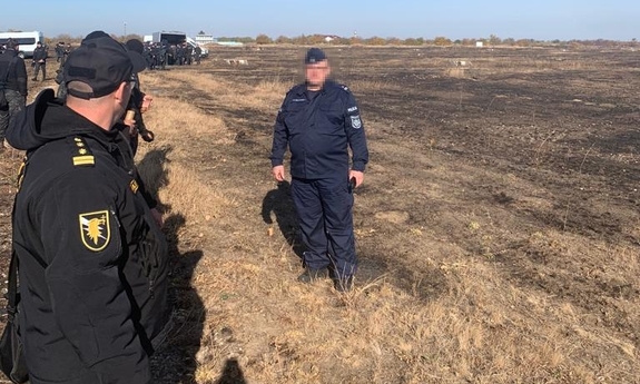 Otwarta przestrzeń. Policjant stoi naprzeciwko funkcjonariuszy stojących w rzędzie.