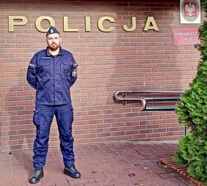 Na tle budynku stoi policjant. Na budynku napis POLICJA