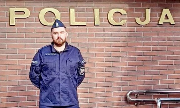 Na tle budynku stoi policjant. Na budynku napis POLICJA