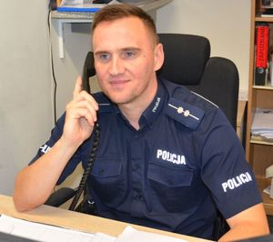 umundurowany policjant siedzi przy biurku, w ręku trzyma słuchawkę telefonu