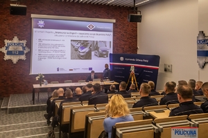 policjanci i inne osoby siedzące na widowni auli podczas konferencji, na ekranie wyświetlana jest prezentacja