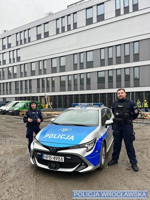 Przy radiowozie, na tle budynków, stoi dwóch policjantów