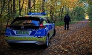 Radiowóz w lesie a przed nim stojący policjant patrzący w głąb lasu