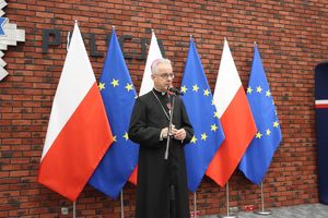 Ksiądz w czarnej sutannie stoi na tle flag Polski i Unii Europejskiej