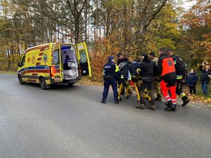 Na asfaltowej drodze stoi żółta karetka. Policjanci i ratownicy medyczni na specjalnym łóżku na kołach wiozą pacjenta