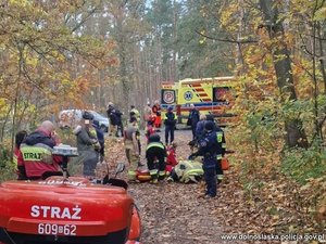 Na leśnej drodze stoją policjanci i strażacy. Po prawej stronie karetka. Po lewej motocykl strażacki