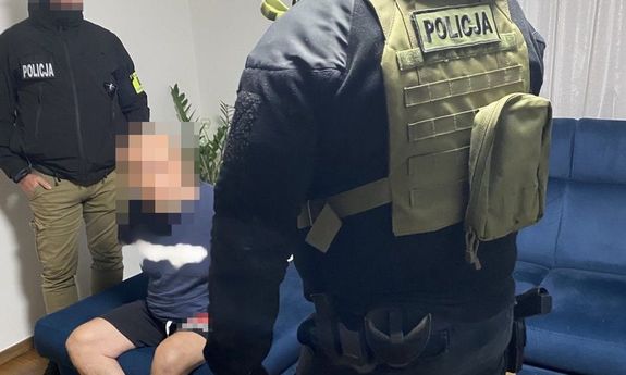 Dwóch policjantów stoi przy zatrzymanym mężczyźnie, który siedzi na niebeskiej kanapie