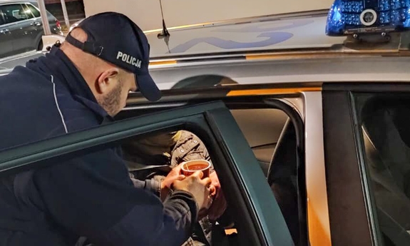 umundurowany policjant podaje herbatę mężczyźnie siedzącemu w radiowozie