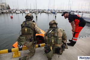 Na moście przy wodzie stoją policyjni kontrterroryści i ratownik medyczny. Na wodzie pomarańczowy pływający ponton w kształcie trójkąta