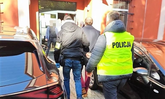 na zdjęciu widać nieumundurowanych policjantów ubranych w kamizelki z napisem Policja, którzy prowadzą zatrzymanego mężczyznę do budynku prokuratury