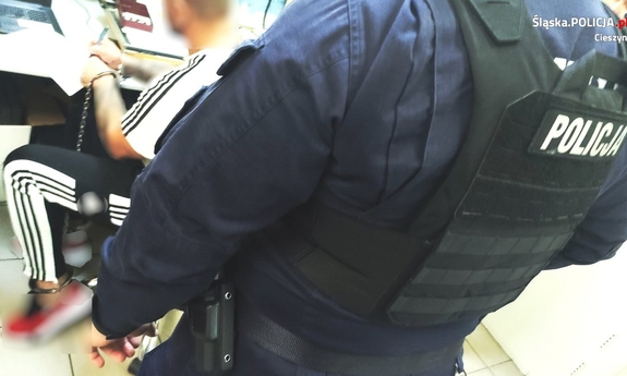 policjant w mundurze pilnuje zatrzymanego mężczyzny. Zatrzymany ma kajdanki założone na ręce i nogi