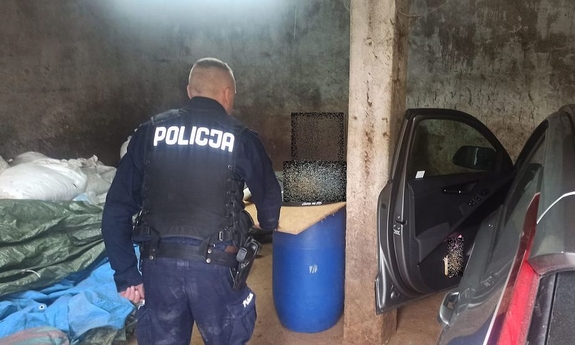 W garażu stoi zaparkowany samochód, , leża jutowe worki obok stoi umundurowany policjant