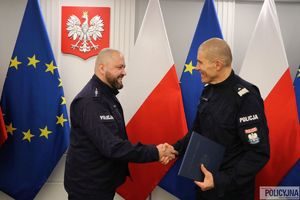 Zastępca Komendanta Głównego Policji nadinsp. Roman Kuster gratuje policjantowi. W tle flagi Polski i Unii Europejskiej w stojakach flagowych, na ścianie wisi godło Polski.