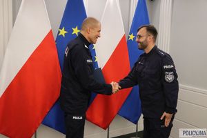 Zastępca Komendanta Głównego Policji nadinsp. Roman Kuster gratuje policjantowi. W tle flagi Polski i Unii Europejskiej w stojakach flagowych, na ścianie wisi godło Polski.