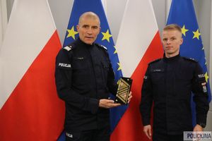 Zastępca Komendanta Głównego Policji nadinsp. Roman Kuster z policjantem na tle flag Polski i Unii Europejskiej w stojakach flagowych, na ścianie wisi godło Polski.