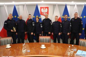 Zastępca Komendanta Głównego Policji nadinsp. Roman Kuster z policjantami na tle flag Polski i Unii Europejskiej w stojakach flagowych, na ścianie wisi godło Polski.