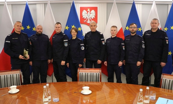 Zastępca Komendanta Głównego Policji nadinsp. Roman Kuster z policjantami na tle flag Polski i Unii Europejskiej w stojakach flagowych, na ścianie wisi godło Polski