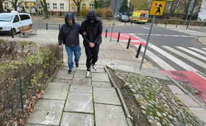 Nieumundurowany policjant prowadzi chodnikiem zatrzymanego mężczyznę zakutego w kajdanki