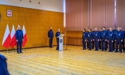 przy mównicy stoi policjantka przemawia do mikrofonu, za nią przy ścianie po lewej i prawej stronie stoją dwaj policjanci, za policjantem z lewej strony stoją cztery flagi Polski w stojakach