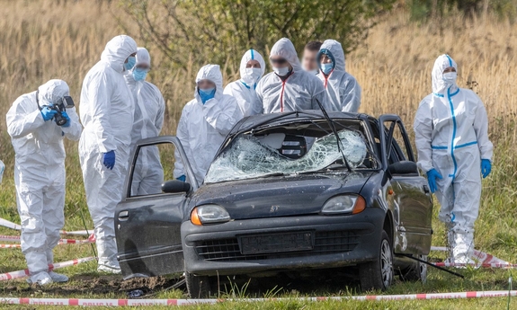 ubrani w białe kombinezony funkcjonariusze  stoją przy czarnym rozbitym samochodzie