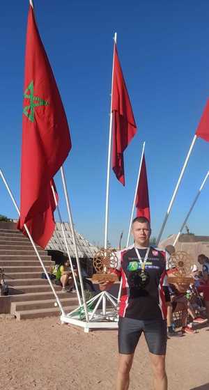 Asp. Marcin Brewczyński w stroju sportowym ze statuetkami w dłoniach stoi na tle marokańskich flag znajdujących się w stojaku