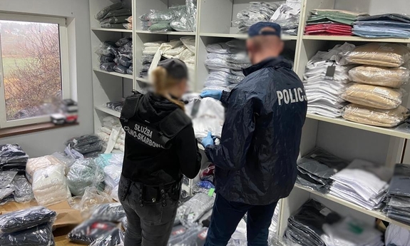 Funkcjonariusze przeszukują pomieszczenie, na regałach leżą ubrania zapakowane w foliowe worki