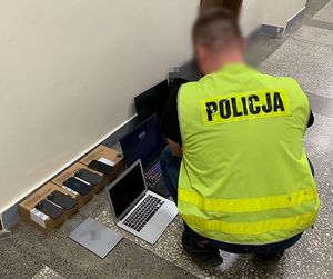 policjant kuca obok zabezpieczonego sprzętu, laptopów i telefonów komórkowych