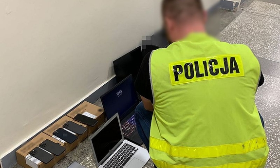 policjant kuca obok zabezpieczonego sprzętu, laptopów i telefonów komórkowych