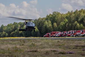 śmigłowiec wznosi się nisko nad płytą lotniska, w głębi po prawej stronie wozy strażackie