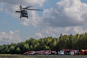 śmigłowiec wznosi się nad płytą lotniska, w głębi po prawej stronie wozy strażackie