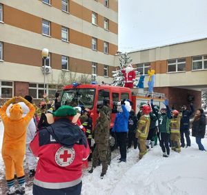 Maskotki, policjanci, wojskowi, strażacy machają dzieciom stojącym w oknach szpitala. Obok stoi wóz strażacki