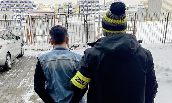 Policjant prowadzi zatrzymanego na policję. Dookoła śnieg. Policjant ma kolorową czapkę i czarną kurtkę