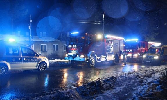 Noc. Przed budynkiem stoją dwa wozy straży pożarnej, radiowóz oraz karetka