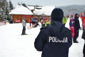 Plecy policjanta i zbliżenie na jego kurtkę z napisem policja. W tle ludzie na stoku