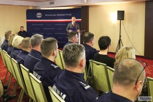 policjanci siedzą podczas konferencji na krzesłach ustawionych w rzędy, w głębi mężczyzna stojący przy mównicy