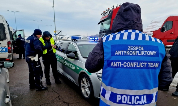 odwrócony tyłem funkcjonariusz zespołu antykonfliktowego, w tle pojazd słuzby celno-skarbowej i funkcjonariusze