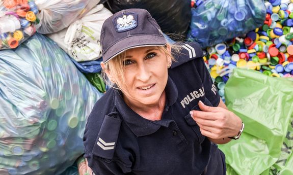 Na pierwszym planie stoi policjantka. W tle leżą worki zapełnione korkami.